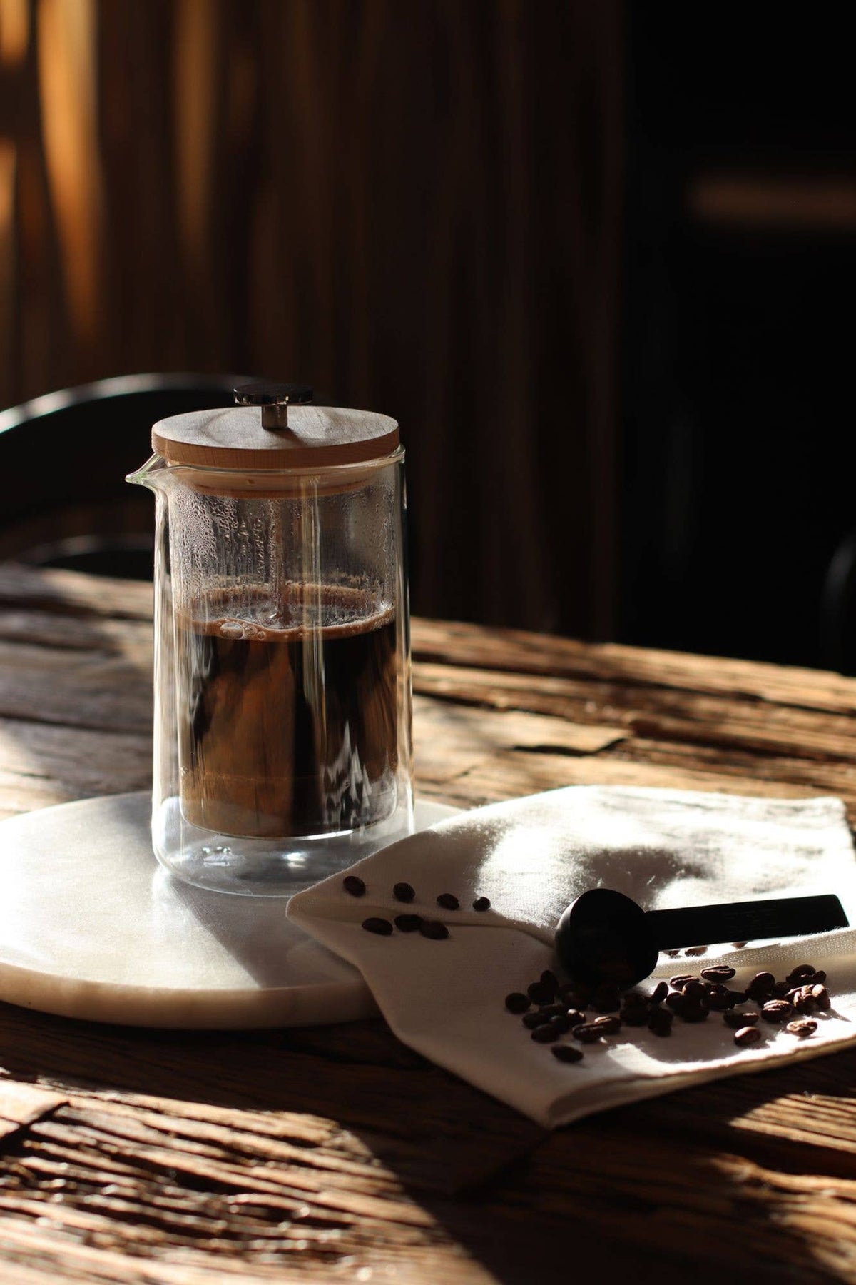 A clear glass French press with a wooden lid sits on a white marble coaster on a rustic wooden table, with coffee inside. A folded white cloth with scattered coffee beans and a black spoon rests beside it in warm, dramatic sunlight.