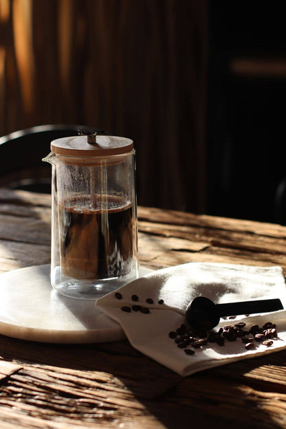 A clear glass French press with a wooden lid sits on a white marble coaster on a rustic wooden table, with coffee inside. A folded white cloth with scattered coffee beans and a black spoon rests beside it in warm, dramatic sunlight.